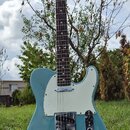 Modře lakovaná elektrická kytara typu Telecaster s bílým pickguardem, umístěná na černém stojanu v trávě. Na hlavě hmatníku je viditelné logo Fender.…