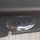 Detail černého plastového povrchu s oválnou stříbrnou nálepkou. Na nálepce je uveden nápis FOCUS a pod ním menším písmem percussion. Povrch plastu vy…