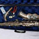 Stříbrný saxofon v černém kufru s modrým polstrováním. Vedle nástroje je položen munční dutník, červený popruh a modré krabičky s příslušenstvím. Na…