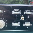 Detail ovládacího panelu černého hudebního přístroje. Panel obsahuje bílý přepínač s nápisem Instr. 2, čtyři posuvníky označené symboly houslového a…