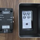 Na fotografii jsou dvě zařízení na dřevěném povrchu. Vlevo je černý efektový pedál s bílým logem Laney a nápisy EXPRESSION, INPUT, SIGNAL, OUTPUT, RI…