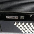 Detail černého akordeonu s bílými knoflíky. Na černé ploše je umístěn stříbrný nápis CHORAL a vedle něj malý štítek s označením VI. Povrch nástroje v…