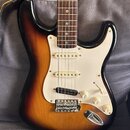 Elektrická kytara s tmelem v barvě sunburst a bílým pickguardem. Nástroj má tři snímače typu single-coil, chromovaný most a chromovanou páčku vibrata…