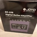 Černý karton s produktem JOYO DC-15S Digital Guitar Amplifier. Na krabici je bílé logo JOYO s nápisem FEEL JOYO ENJOY MUSIC a fotografie zadní strany…