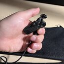 Ruka drží malý černý plastový klip s integrovaným mikrofonem. Zařízení je připojeno k tenkému černému kabelu. Povrch plastu vykazuje drobné známky op…