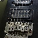 Černá elektrická kytara s hnedým hmatníkem. Nástroj má tři snímače, z nichuž spodní je humbucker. Je zde instalován kovový vibrační systém s šesti la…