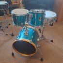 Modrázlatý glitrový bicí set skládající se z basového bubnu, dvou tomů a samostatného snare drumu na stojanu. Na jednom z tomů je viditelné kulaté lo…