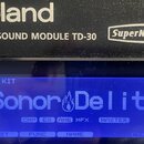 Detailní záběr na horní část a displej elektronického bubnového modulu. Na černém povrchu je bílý nápis Roland, DRUM SOUND MODULE TD-30 a logo SuperN…
