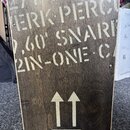 Detail dřevěného nástroje s tmavým povrchem a světlými nápisy v provedení šablony. Na horní části je text „HERK PERCU“, „2,60' SNARI“ a „2IN-ONE C/“.…