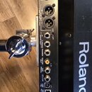 Detail zadní strany černého elektronického zařízení s bílým nápisem Roland. Jsou zde vidět různé konektory a popisky: INPUT BASS, OUTPUT, R SUB OUT (…