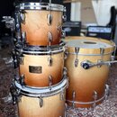 Sada bubnů v oranžovém přechodovém odstínu s viditelnou strukturou dřeva. Na prostředním tomu je kovový štítek s nápisem Pearl Masters Custom. Horní…