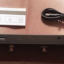 Černý obdélníkový přístroj s čtyřmi kovovými konektory na spodní straně. Na zadním panelu jsou vidět tři porty: napájecí vstup, USB-C a USB-A. Vedle…