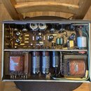 Fotografie zachycuje vnitřek elektronického zařízení, pravděpodobně zesilovače, s otevřeným šasi. Jsou zde vidět tři elektronky, kondenzátory a dva t…