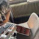 Detailní pohled na hřif a tělo akustické kytary s tmavým dřevěným povrchem. Na těle jsou vidět tři černé ovládací knoflíky. V pozadí je šedá židle, b…