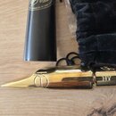 Zlatý kovový mouthpiece s logem ve tvaru kruhu s přínosným prvkem. Na nátrubu je vyryté označení NY a nad ním nečitelný podpis. Vedle nástroje je čer…