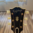 Fotografie zachycuje hlavu elektrické kytary v černé barvě s bílým lemováním. V horní části je bílý nápis Epiphone a uprostřed se nachází bílé logo v…