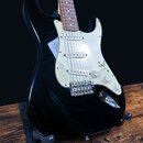 Černá elektrická kytara typu Stratocaster s bílým pickguardem a hnedým hmatníkem. Na ovládacích knoflících jsou nápisy VOLUME a TONE. Nástroj vykazuj…