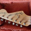 Fotografie zachycuje hlavu elektrické kytary v přírodním dřevěném odstínu, která leží v červeném plyšovém pouzdře. Na hlavě je vidět logo Fender, náp…