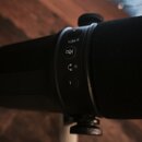 Detailní záběr na černý mikrofon s černé molitovou clonou. Na těle mikrofonu jsou vidět ovládací tlačítka s ikonami pro přičítání (+), ztlumení zvuku…