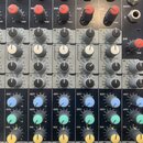Detailní pohled na ovládací panel míchacího pultu s několika kanály. Jsou vidět řady šedých a červených otočných knoflíků s nápisy GAIN, HF, MF, LF,…