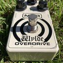 Bílý efekt pedál s nápisy Wylde OVERDRIVE a logem MXR. Na horní straně jsou tři černé ovládací knoflíky s popisky OUTPUT, TONE a GAIN. Na šasi jsou v…