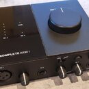 Černý audio rozhraní s nápisem KOMPLETE AUDIO 1 a logem výrobce. Na horní části jsou popisky IN 1, IN 2, USB a 48 V u kontrolních světel, vpravo je v…