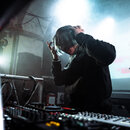 Fotografie zachicuje osobu v černé mikině za DJským pultem. V popředí je vidět mixážní pult s mnoha knoflíky a posuvníky, vedle kterého leží černá DJ…
