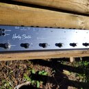 Stříbrný kovový pedálboard s šesti černými spínači. Na povrchu je nápis „Harley Benton“ a označení „STOMPISOLE 4 ISO“. Jsou zde dále nápisy „DC IN“,…