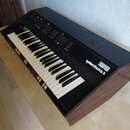 Černý elektronický klávesový nástroj s dřevěnými bočními stěnami. Na pravé straně je bílý nápis FARFISA a pod ním menší nápis syntorchestra 4. Horní…