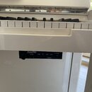 Detail bílého klavíru s viditelnou klávesnicí. Pod klávesnicí je umístěn černý ovládací panel s nápisem PianoDisc. Panel obsahuje ovratový knoflík VO…