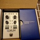 Bílý elektronický přístroj s nápisem SDRUM Strummable Drums. Obsahuje dva otočné ovladače, několik tlačítek s popisky jako KICK a SNARE a různé přepí…