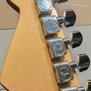 Detail hlavy elektrické kytary z přírodního dřeva s šesti chromovanými ladičkami. Na každém krytu ladičky je vyryto logo „Fender“. Povrch dřeva vykaz…