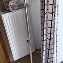 Stříbrný kovový mikrofonní stojan s trojnohou základnou. Stojan má teleskopický mechanismus s dvěma utahovacími šrouby. Na horní části je vidět držák…
