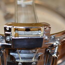 Detailní záběr na chromovaný mechanismus snare thrower u bubnu. Kovový povrch vykazuje drobné škrábance a známky opotřebení. Na fotce nejsou žádné vi…