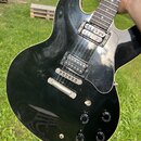 Černá elektrická kytara s tmavým hnedým hmatníkem a světle béžovým lemováním korpusu. Nástroj má dva humbuckery, čtyři ovládací knofíky a černý plech…