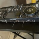 Černý DJ 컨트롤러 s dvěma jogovými koly a centrálním displejem. Na přední straně je bílý nápis Numark MIXSTREAM PRO a logo engresos. Zadní panel obsahuje…