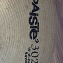Detailní záběr na povrch činchy v přirozené zlatavé barvě s viditelným opracováním povrchu. Na činchy je černý nápis PAISTE 302 a pod ním text 14" Hi…
