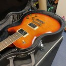 Elektrická kytara s oranžovo-žlutým gradientem v černé poloměkkejem pouzdře. Nástroj má dva humbuckery, čtyři zlaté ovládací knoflíky a zlatý most. N…