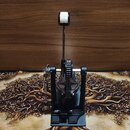 Fotografie zobrazuje černý pedál basového bubnu s kovovou konstrukcí a bílým šlapadlem. Na zařízení nejsou vidět žádné značky, loga ani modelové ozna…