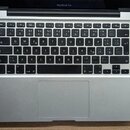 Fotografie znázorňuje otevřený notebook stříbrné barvy s černou klávesnicí a trackpadem. Na horním rámu displeje je nápis MacBook Pro. Zařízení je um…