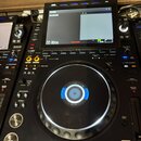 Černý DJ mixer s displejem, na kterém je nápis SOURCE NO DEVICE a Not Loaded. V horní části jsou modré tlačítka, ve spodní části je velký jog wheel s…