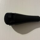 Černý mikrofon s nasazenou černou pěnovou windscreen. Tělo mikrofonu je z matního černého materiálu s mřížkou v horní části. Na zařízení nejsou vidit…