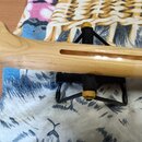Detail krku kytary z přírodního světlého dřeva, která je umístěna na černém skládacím stojanu. V horní části krku je viditelný podélný výřez (slot).…