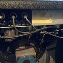 Detail zadního panelu zesilovače s černým povrchem a čtyřmi vstupy. Nad konektory jsou nápisy VIBRATO PEDAL, REVERB PEDAL, REVERB L a REVERB IN. Pod…