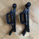 Dvě černé kovové stojany na nástroje s polstrovanými rukojtěmi. Stojany jsou umístěny vedle sebe na dřevěné podlaze. Na předmětech nejsou viditelné ž…