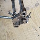 Fotografie zachycuje detail chromovaného kovového mechanismu, pravděpodobně části stojanu nebo pedálu. Součásti jsou spojeny šrouby s křídlovými mati…