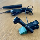 Černý mikrofon s připevněnou svorkou a zeleným gumovým odpružňovačem. Mikrofon je připojen k dlouhému černému kabelu, který končí v černém válcovém m…