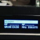 Černý bezdrátový přijímač s kovovými stříbrnými prvky na bocích. Na modrém displeji je text MODER:1, RF ----, AF ----, GR.08 CH.08 MHz 598.875. Na pa…