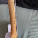 Detailní pohled na hmatník a krk elektrické kytary z profilu. Krk je vyroben ze světlého dřeva s lakovaným povrchem, tělo kytary v pozadí je bílé. Na…