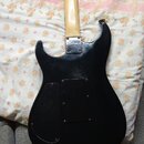 Zadní strana černé elektrické kytary s jasným kytarovým hředem. Na zadní straně těla je kovový kryt s nápisem "Squier by Fender". Povrch laku vykazuj…